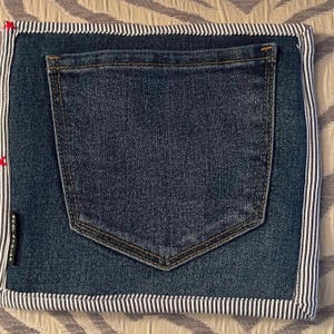 May include: A denim-style pouch with a dark blue jean pocket sewn onto the front. The edges are trimmed with a white and navy blue striped fabric. The left side has a white label with the text "WHATATRIPP".