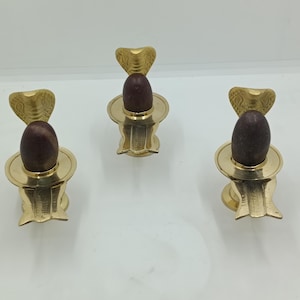 May include: Three brass Shiva Lingam statues. Each statue features a brown stone lingam atop a brass base with a cobra hood design. The statues are displayed on a white surface.