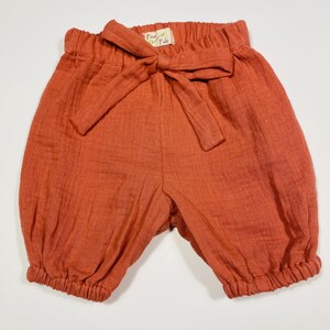 Könnte beinhalten: Rostfarbene Babyshorts mit einer Schleife an der Taille. Die Shorts haben einen elastischen Bund und geraffte Bündchen. Auf dem Etikett der Schleife steht "Pine & Pale". Die Shorts sind aus einem strukturierten Stoff gefertigt.