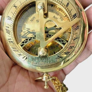 Puede incluir: Una br&uacute;jula de sol de lat&oacute;n vintage con n&uacute;meros romanos y una cadena. La br&uacute;jula presenta una esfera detallada con marcas direccionales y una tapa con bisagras. El texto "F.L. West 91, Look Spur, London" es visible.