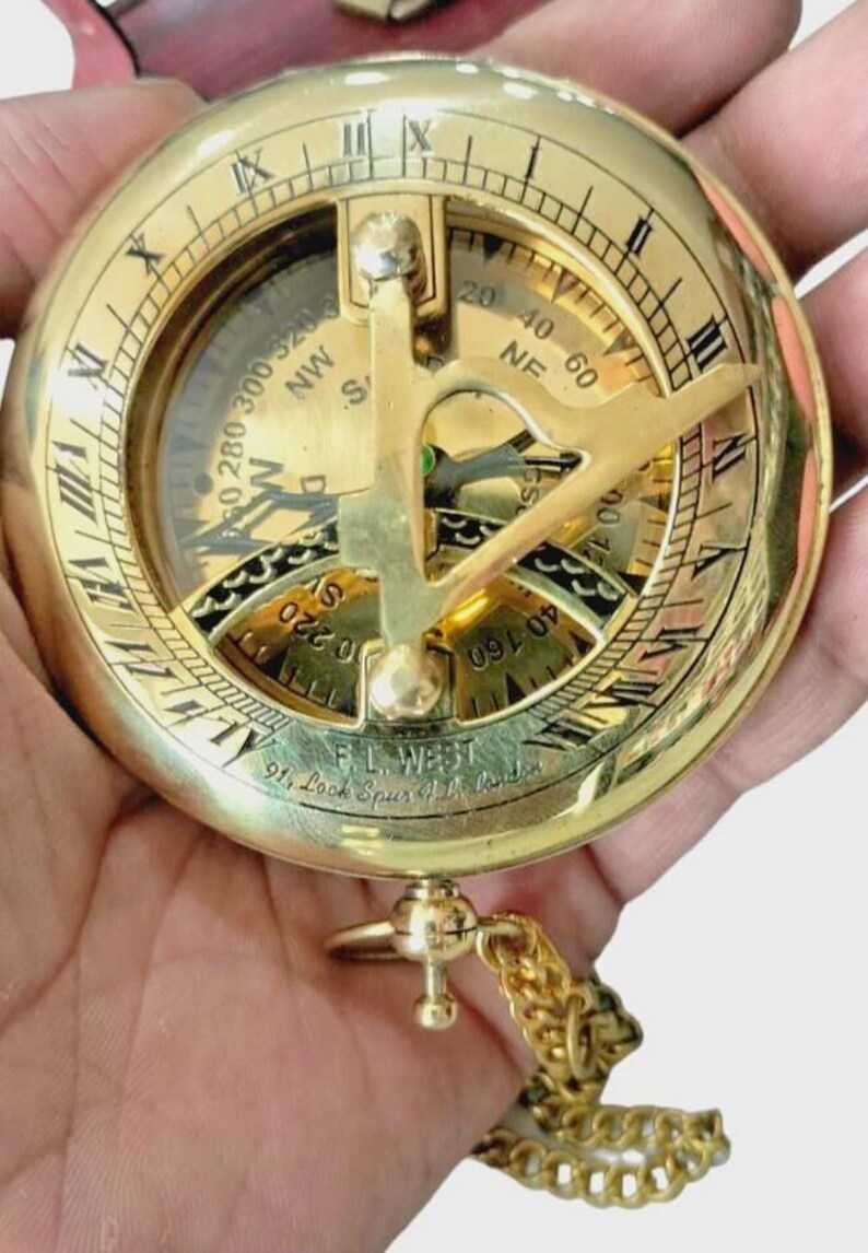 Puede incluir: Una br&uacute;jula de sol de lat&oacute;n vintage con n&uacute;meros romanos y una cadena. La br&uacute;jula presenta una esfera detallada con marcas direccionales y una tapa con bisagras. El texto "F.L. West 91, Look Spur, London" es visible.