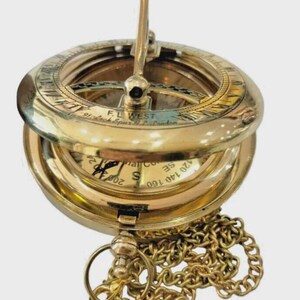 Puede incluir: Una br&uacute;jula de reloj de sol de lat&oacute;n vintage con tapa con bisagras y cadena. La br&uacute;jula presenta marcas grabadas y un gnomon. El instrumento se utiliza para determinar la hora del d&iacute;a.