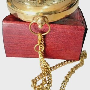 Puede incluir: Una br&uacute;jula de sol de lat&oacute;n vintage con una cadena dorada, sobre una caja de madera roja. La br&uacute;jula presenta marcas grabadas y la inscripci&oacute;n "F.L. WEST, Bush Spurs, London."