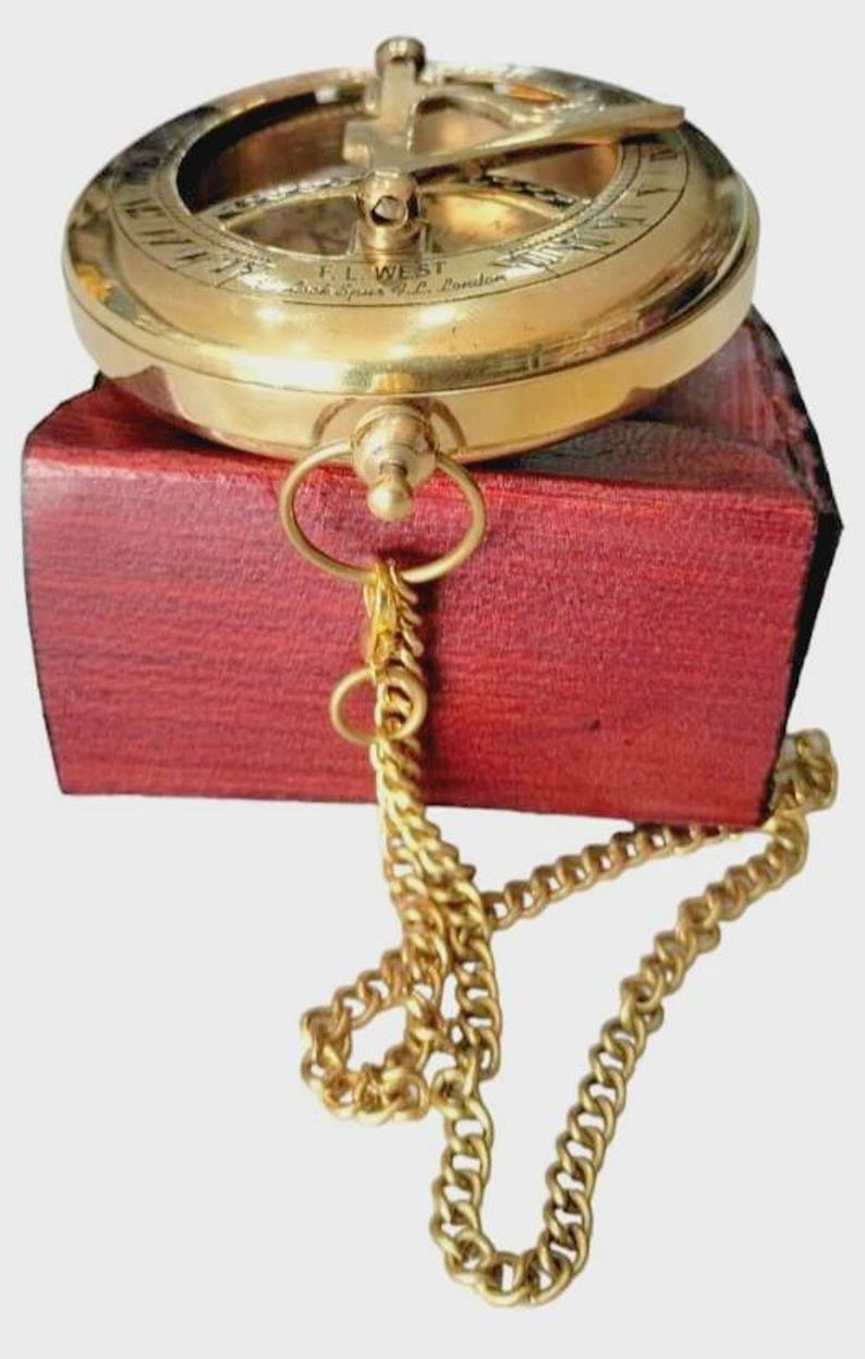Puede incluir: Una br&uacute;jula de sol de lat&oacute;n vintage con una cadena dorada, sobre una caja de madera roja. La br&uacute;jula presenta marcas grabadas y la inscripci&oacute;n "F.L. WEST, Bush Spurs, London."