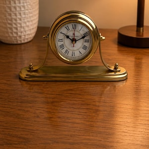 May include: A polished brass mantel clock featuring a circular face with black Roman numerals and thin black hands. The clock is mounted on a curved brass stand with a rectangular base, showcasing a classic design with a white dial background.