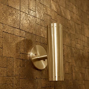 May include: A cylindrical wall sconce with a brushed metallic finish, featuring a series of small circular perforations along the top and bottom edges. The fixture is mounted to a circular base plate, providing a clean and functional lighting design.