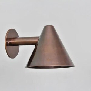 May include: A bronze-colored wall-mounted light fixture. The fixture has a circular base, a cylindrical arm, and a conical shade. The light is designed to be mounted on a wall and provides directional lighting. The fixture is made of metal.