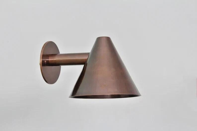 May include: A bronze-colored wall-mounted light fixture. The fixture has a circular base, a cylindrical arm, and a conical shade. The light is designed to be mounted on a wall and provides directional lighting. The fixture is made of metal.