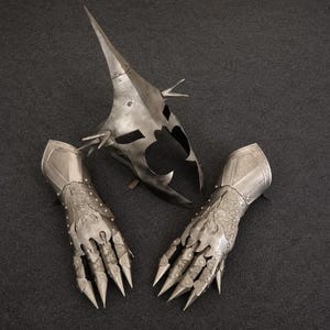 May include: A silver-toned metal helmet featuring a tall, pointed crown and side spikes, accompanied by a pair of matching articulated gauntlets. The gauntlets display intricate etched patterns and long, sharp, claw-like finger plates.