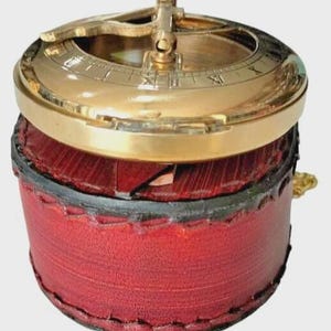 Puede incluir: Un reloj de sol de lat&oacute;n antiguo con n&uacute;meros romanos, colocado sobre una caja cil&iacute;ndrica de cuero rojo. El reloj de sol presenta un gnomon dorado y una esfera circular. La caja est&aacute; cosida con un borde negro, creando una est&eacute;tica cl&aacute;sica y antigua.