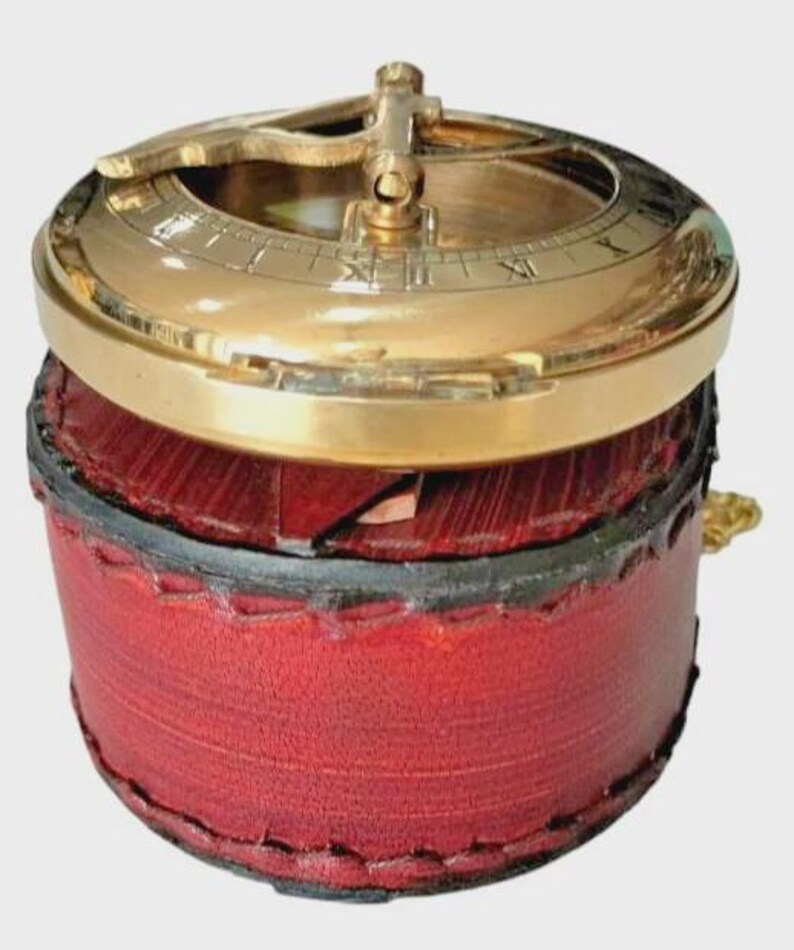 Puede incluir: Un reloj de sol de lat&oacute;n antiguo con n&uacute;meros romanos, colocado sobre una caja cil&iacute;ndrica de cuero rojo. El reloj de sol presenta un gnomon dorado y una esfera circular. La caja est&aacute; cosida con un borde negro, creando una est&eacute;tica cl&aacute;sica y antigua.