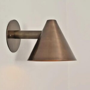 May include: A bronze-colored wall sconce with a conical shade and a circular backplate. The sconce has a cylindrical arm connecting the shade to the backplate. The fixture is mounted on a white wall, providing a minimalist aesthetic.