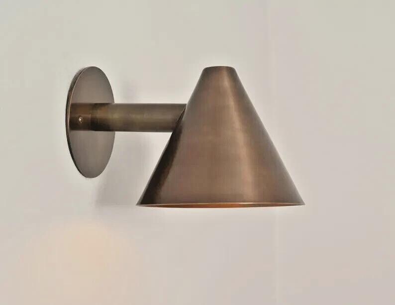 May include: A bronze-colored wall sconce with a conical shade and a circular backplate. The sconce has a cylindrical arm connecting the shade to the backplate. The fixture is mounted on a white wall, providing a minimalist aesthetic.