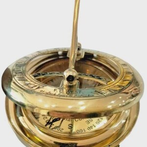 Puede incluir: Un reloj de sol de lat&oacute;n vintage con tapa abatible y cadena. El reloj de sol presenta un dise&ntilde;o circular con n&uacute;meros romanos e indicaciones direccionales. El texto "F.L. WEST 91, Lock Spur & Co. London" es visible.