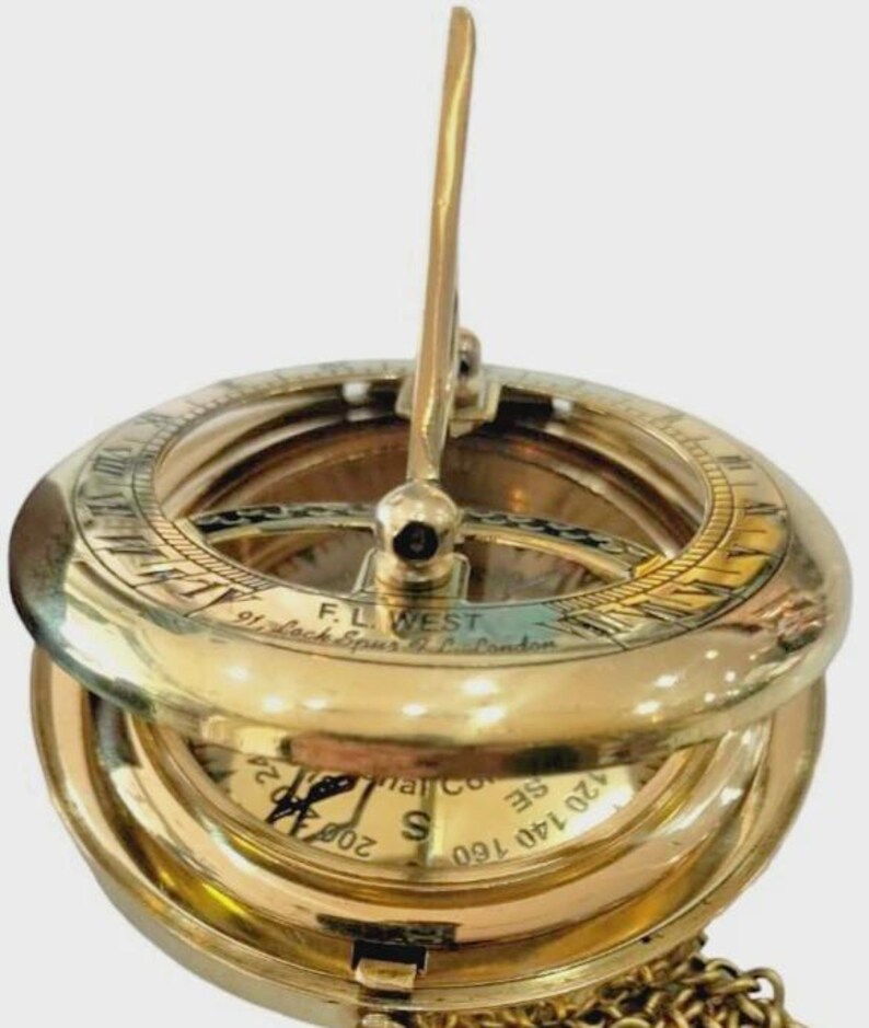 Puede incluir: Un reloj de sol de lat&oacute;n vintage con tapa abatible y cadena. El reloj de sol presenta un dise&ntilde;o circular con n&uacute;meros romanos e indicaciones direccionales. El texto "F.L. WEST 91, Lock Spur & Co. London" es visible.