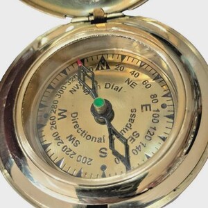 Puede incluir: Una br&uacute;jula de lat&oacute;n vintage con tapa con bisagras y cadena. La esfera de la br&uacute;jula presenta marcas direccionales, una aguja y las palabras "Directional Compass" y "Dial". La br&uacute;jula es una herramienta de navegaci&oacute;n cl&aacute;sica.