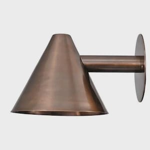 May include: A bronze-colored wall sconce with a conical shade and a cylindrical arm. The sconce has a circular mounting plate. The metal fixture is designed for indoor or outdoor lighting.