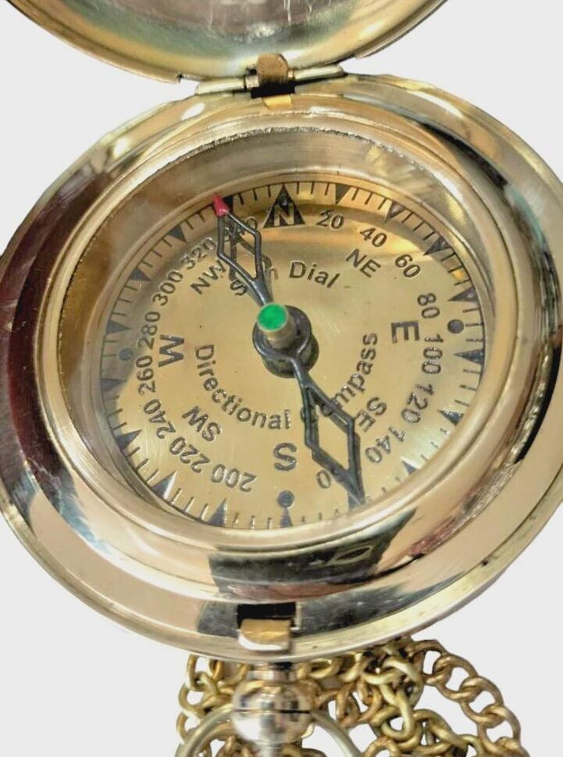 Puede incluir: Una br&uacute;jula de lat&oacute;n vintage con tapa con bisagras y cadena. La esfera de la br&uacute;jula presenta marcas direccionales, indicadores de grados y la palabra "Dial". La aguja apunta hacia el norte. Una herramienta de navegaci&oacute;n cl&aacute;sica.