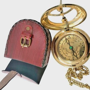 Puede incluir: Una br&uacute;jula de sol de lat&oacute;n vintage con una funda de cuero. La br&uacute;jula presenta una esfera detallada con marcas direccionales y una cadena. La funda de cuero rojo tiene un cierre de hebilla. Una herramienta de navegaci&oacute;n cl&aacute;sica.