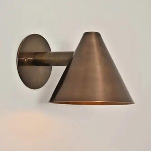 May include: A bronze-colored wall sconce with a conical shade and a circular backplate. The sconce extends from the wall with a short arm. The fixture is made of metal and has a warm, metallic finish. The design is minimalist and modern.