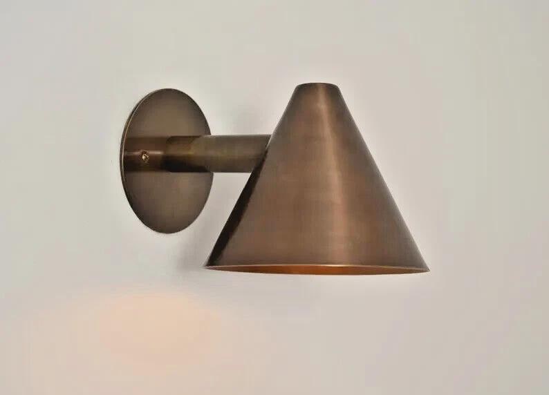 May include: A bronze-colored wall sconce with a conical shade and a circular backplate. The sconce extends from the wall with a short arm. The fixture is made of metal and has a warm, metallic finish. The design is minimalist and modern.
