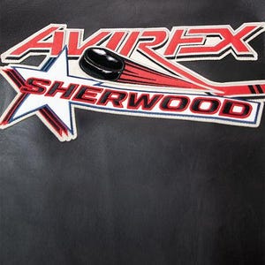 May include: Black leather jacket with a hockey-themed patch. The patch features the word "AVIREX" in red and white, a hockey puck, and the word "SHERWOOD" in red and white. A small logo is at the top.