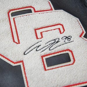 May include: Close-up of a white and red letterman jacket patch with the letters "GB" and a signature. The letters are white with a red outline, and the background is black.