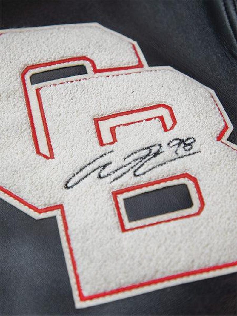 May include: Close-up of a white and red letterman jacket patch with the letters "GB" and a signature. The letters are white with a red outline, and the background is black.