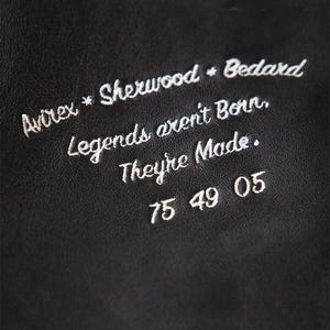 May include: Close-up of a black leather surface with white embroidered text. The text includes the words "Avirex * Sherwood * Bedard" and the phrase "Legends aren't Born, They're Made." Below the text are the numbers "75 49 05".