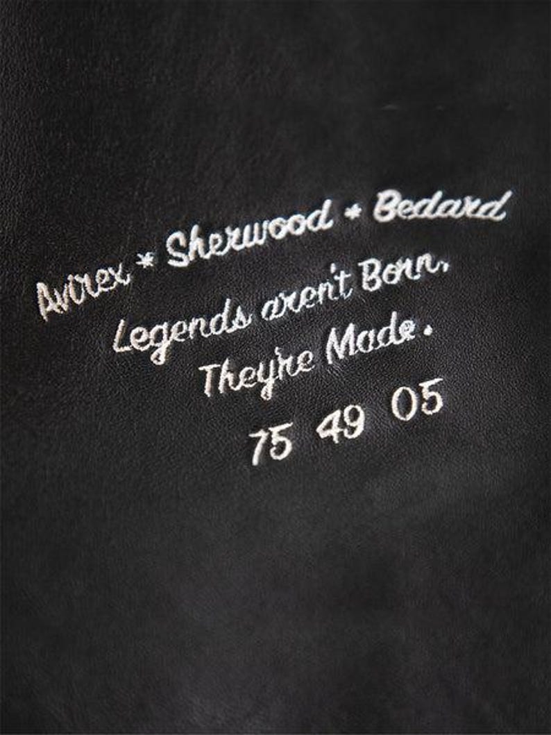 May include: Close-up of a black leather surface with white embroidered text. The text includes the words "Avirex * Sherwood * Bedard" and the phrase "Legends aren't Born, They're Made." Below the text are the numbers "75 49 05".