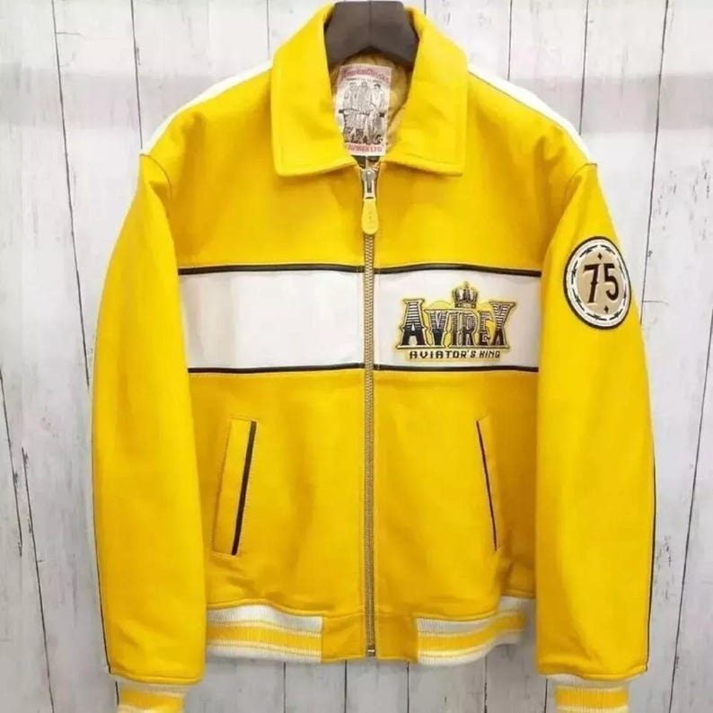 May include: A yellow leather jacket with a white stripe across the chest and sleeves. The jacket features the "AVIREX AVIATOR & KING" logo and a "75" patch. It has a zipper closure and a collar.