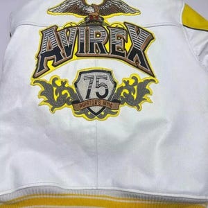 May include: White leather jacket with yellow and gray accents. Features the word "AVIREX" in large, bold lettering, and a patch that says "75 AVIATOR'S KING". An eagle is embroidered above the brand name.