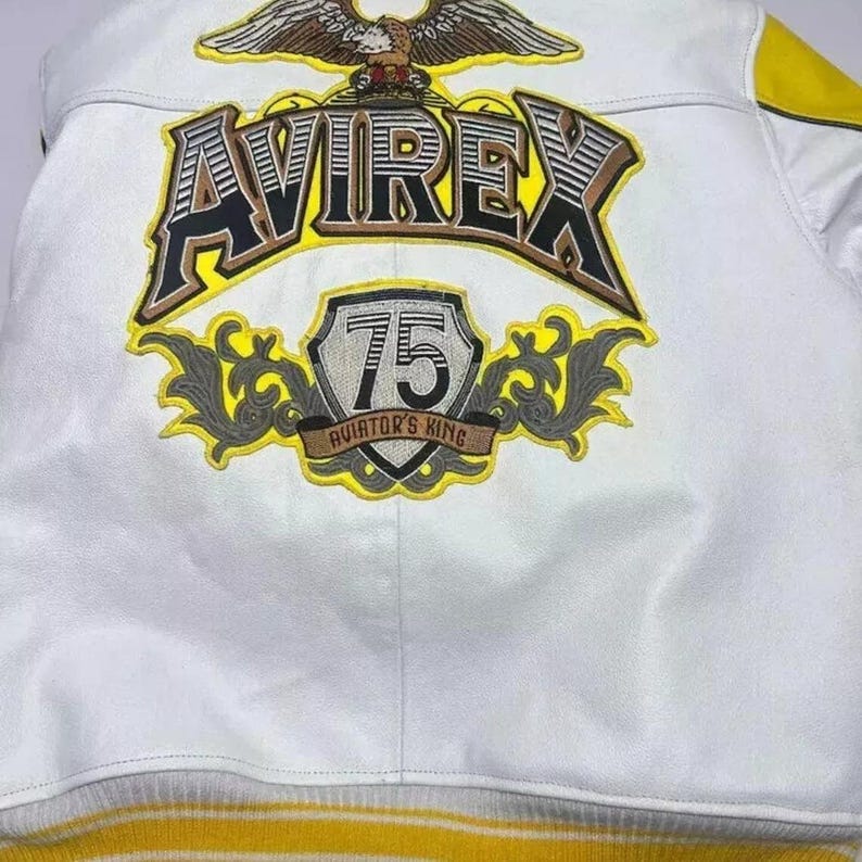 May include: White leather jacket with yellow and gray accents. Features the word "AVIREX" in large, bold lettering, and a patch that says "75 AVIATOR'S KING". An eagle is embroidered above the brand name.