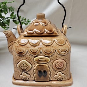 May include: A brown ceramic teapot shaped like a house, featuring a decorative roof and embossed details. It has a black metal handle with a coil grip and a spout for pouring. The teapot is a unique kitchenware item.