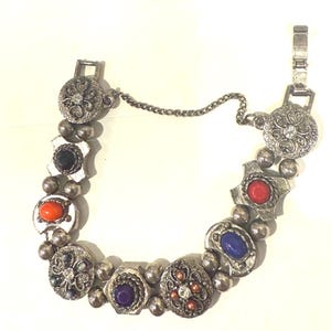May include: Silver-toned bracelet featuring a variety of colorful gemstones and decorative elements. The bracelet includes round, oval, and geometric shapes with red, orange, blue, and purple stones. The clasp and chain are also silver.