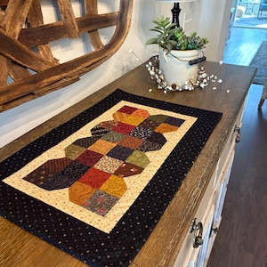 May include: A rectangular quilted table runner featuring a central patchwork design of octagonal shapes in various autumn-toned fabrics including red, gold, brown, and green, surrounded by a cream-colored border and a wide black outer border.