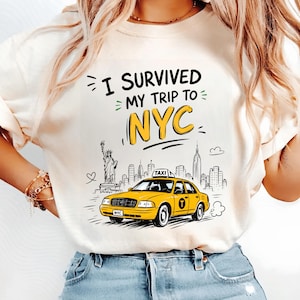 May include: A cream-colored t-shirt featuring a graphic print with the text I SURVIVED MY TRIP TO NYC in black and yellow. The design includes a yellow taxi cab, the Statue of Liberty, and a skyline silhouette of New York City buildings.