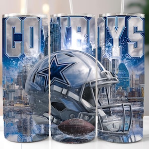 Pode incluir: Um copo de a&ccedil;o inoxid&aacute;vel com o tema Dallas Cowboys. O design inclui a palavra "COWBOYS" em letras prateadas, um capacete de futebol com o logotipo de uma estrela e uma bola de futebol. O fundo mostra uma paisagem urbana.