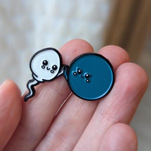 Könnte beinhalten: Ein Emaille-Pin mit einem weißen, spermienförmigen Design mit schwarzer Umrandung und einem lächelnden Gesicht, das einen blauen, kreisförmigen Pin mit einem lächelnden Gesicht umarmt. Die Pins haben einen schwarzen Rand.