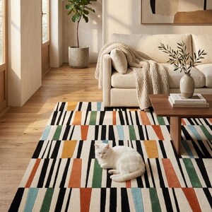 May include: A rectangular area rug featuring a geometric pattern of vertical black stripes interspersed with solid blocks of orange, green, and light blue on a cream background, with a white cat resting in the center of the rug surface.