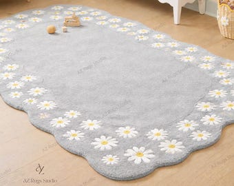 May include: A gray, daisy-shaped rug with a scalloped edge. The rug features embroidered white daisies with yellow centers along the border. The rug is placed on a wooden floor in a room with white furniture.