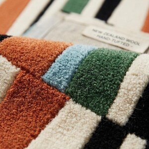 May include: A hand-tufted rug made of New Zealand wool featuring a geometric pattern of rectangular blocks in shades of burnt orange, light blue, forest green, cream, and black. A fabric label reads NEW ZEALAND WOOL - HAND-TUFTED -.