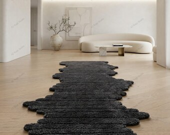 May include: A long, dark charcoal gray runner rug with an irregular, organic, and wavy edge design. The rug features a thick, textured, and tufted pile surface that extends across the light-colored wooden floor in a living room setting.