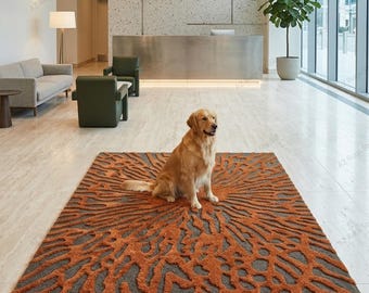May include: A rectangular area rug featuring a textured, raised orange pattern resembling organic, branching coral or root structures set against a flat, dark gray background. A golden-colored dog is sitting in the center of the rug.