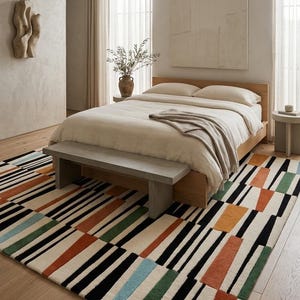 May include: A rectangular area rug featuring a geometric pattern of black, orange, green, and light blue stripes and blocks on a cream-colored background. The rug is positioned on a light wood floor beneath a wooden bed frame and a gray bench.