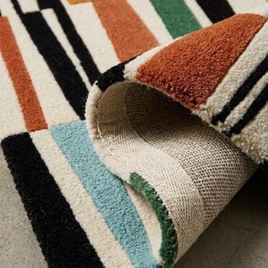 May include: A tufted area rug featuring a geometric pattern of rectangular blocks in black, burnt orange, light blue, and forest green on an off-white background. The rug is folded over to reveal the textured pile and the woven fabric backing.