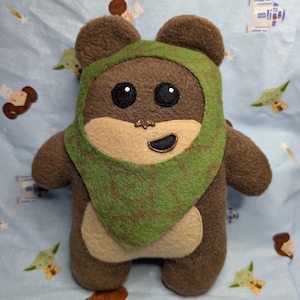 Op de afbeelding: Een handgemaakte Ewok-knuffel. De knuffel heeft een bruin lichaam, een groene capuchon en grote zwarte ogen. De achtergrond is een lichtblauwe stof met Star Wars-personageontwerpen.