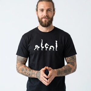 May include: A black short-sleeved t-shirt featuring a white graphic print across the chest. The design displays a sequence of five white silhouettes showing a person performing various yoga poses, including a forward fold, handstand, and bridge.