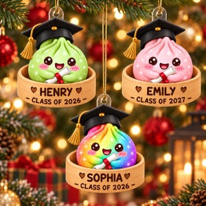 Puede incluir: Tres adornos colgantes con personajes de dumplings con birretes negros y borlas doradas. Cada dumpling sostiene un diploma y se asienta sobre una base de vaporera de bambú grabada con los nombres Henry, Emily o Sophia y años de graduación.