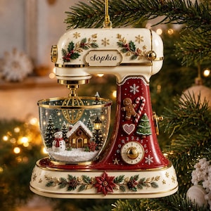 May include: A cream and red stand mixer Christmas ornament featuring a gold whisk and a clear bowl containing a miniature winter scene with a snowman, a house, and trees. The mixer is decorated with holly, snowflakes, and the name Sophia.